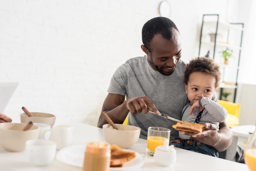 Pai negro segura bebê no colo enquanto passa pasta de amendoim em pão